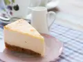 Una fetta di cheesecake con mascarpone, cremosa e invitante, su un piattino rosa. Sullo sfondo, una tazza da tè e una lattiera.