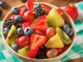 Una colorata macedonia di frutta con fragole, mirtilli, kiwi, ananas e uva, servita in una ciotola su una tovaglia a quadretti verdi.