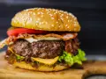 Hamburger fatti in casa con bacon croccante, formaggio filante, pomodoro fresco e lattuga su un tagliere di legno.