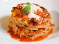 Parmigiana di alici, un piatto stratificato con salsa di pomodoro, formaggio e basilico fresco, servito su un piatto bianco.