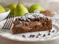 Una fetta di torta pere e cioccolato, cosparsa di zucchero a velo e gocce di cioccolato, con pere caramellate all'interno.