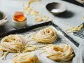 Nidi di pasta fatta in casa su un vassoio infarinato, con uova e una macchina per la pasta sullo sfondo.