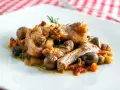 Coniglio alla cacciatora ricetta della nonna: succulenti pezzi di coniglio con olive, pomodorini e rosmarino su un piatto bianco.