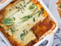 Lasagne di zucchine al forno, dorate e filanti, guarnite con basilico e timo. Una porzione è già servita su un piatto bianco.