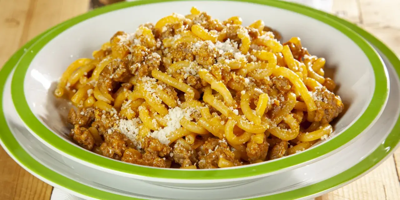 Piatto di gramigna alla salsiccia, condita con ragù e parmigiano grattugiato, servita in una ciotola con bordo verde.