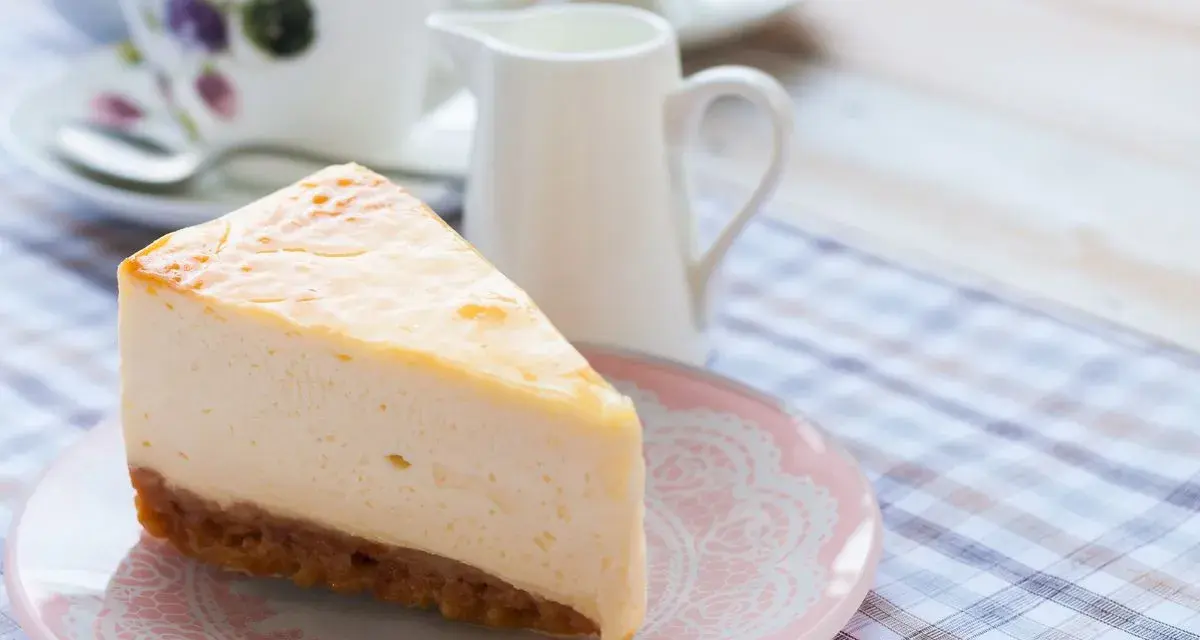 Una fetta di cheesecake con mascarpone, cremosa e invitante, su un piattino rosa. Sullo sfondo, una tazza da tè e una lattiera.
