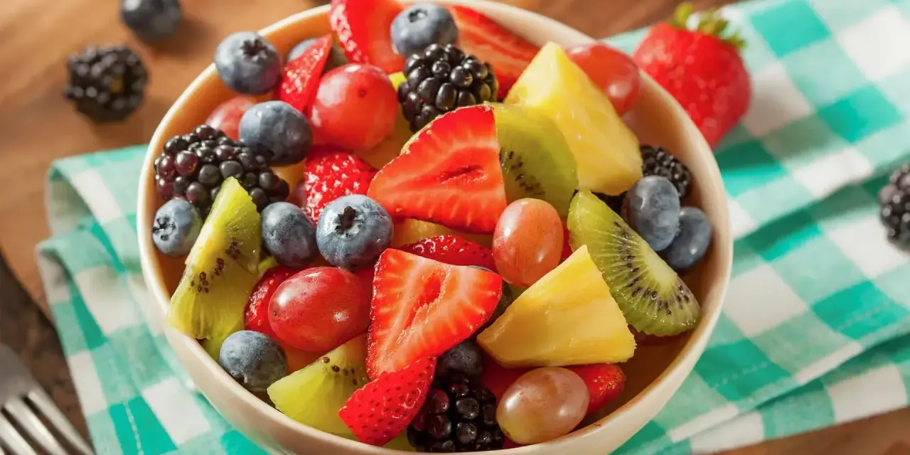 Una colorata macedonia di frutta con fragole, mirtilli, kiwi, ananas e uva, servita in una ciotola su una tovaglia a quadretti verdi.