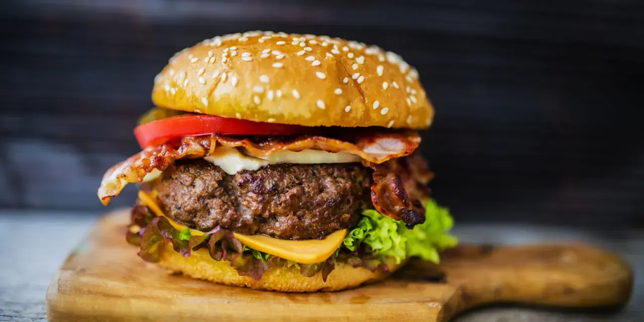 Hamburger fatti in casa con bacon croccante, formaggio filante, pomodoro fresco e lattuga su un tagliere di legno.