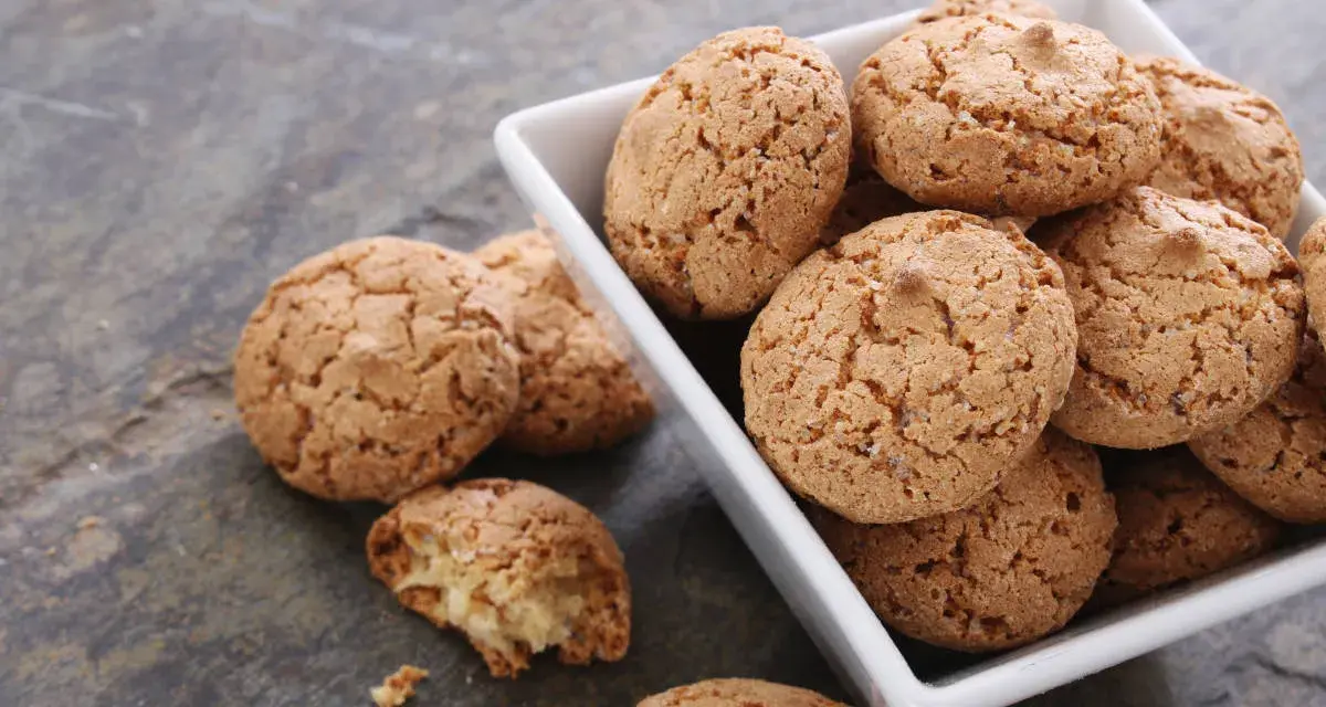 Un mucchio di croccanti biscotti alle mandorle in una ciotola bianca, alcuni sparsi su una superficie grigia.