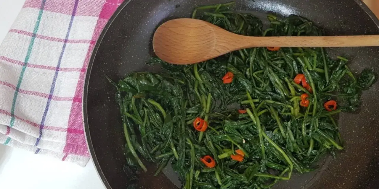 Cicoria in padella con peperoncino, un contorno saporito e salutare. Un cucchiaio di legno mescola le foglie verdi.