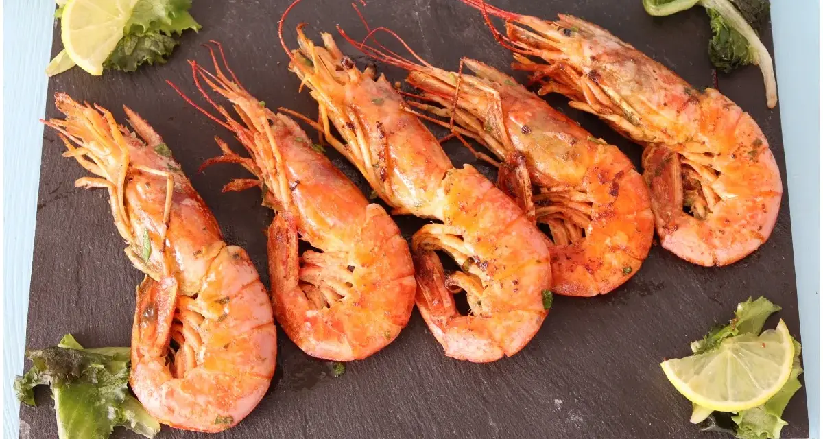 Cinque succulenti gamberoni alla griglia, serviti su una pietra scura con spicchi di limone e insalata fresca.