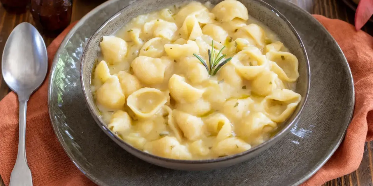Una ciotola di minestra di patate cremosa con pasta a conchiglia, guarnita con rosmarino. Un piatto confortante e saporito.