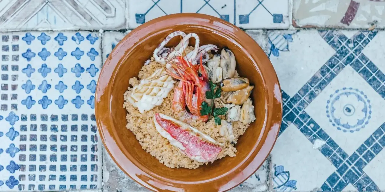 Un invitante cous cous di pesce con gamberi, calamari, cozze e pesce rosso, guarnito con prezzemolo, servito in una ciotola di terracotta su un piatto di maioliche.