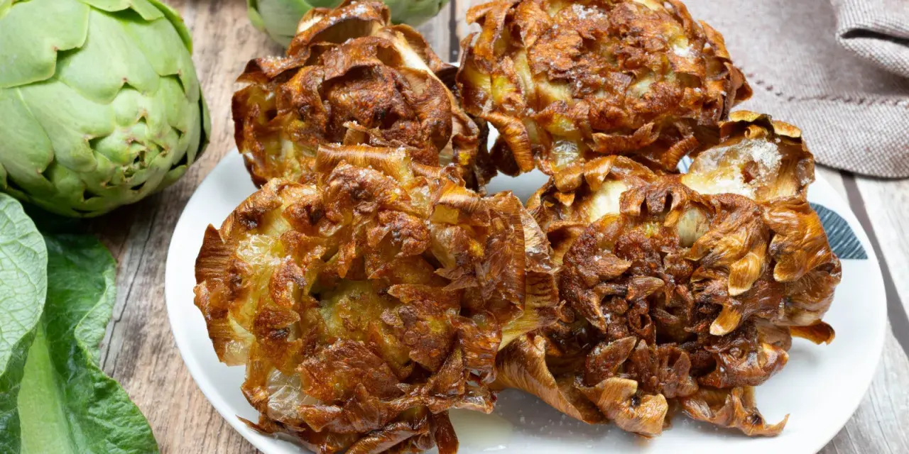 Carciofi alla giudia dorati e croccanti su un piatto bianco, con carciofi freschi sullo sfondo.
