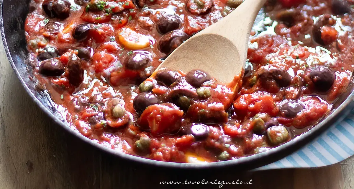 Un cucchiaio di legno raccoglie un sugo ricco e saporito, pronto per condire la pasta alla puttanesca. Pomodoro, olive e capperi si mescolano in un tripudio di sapori mediterranei.