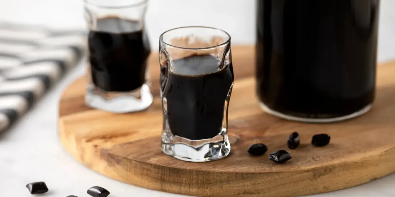 Due bicchierini e una bottiglia di liquore alla liquirizia scuro, con caramelle di liquirizia sparse su un tagliere di legno.
