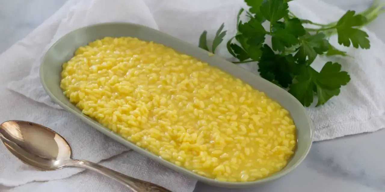 Un cremoso risotto allo zafferano in un piatto ovale, con un cucchiaio d'argento e un rametto di prezzemolo fresco.