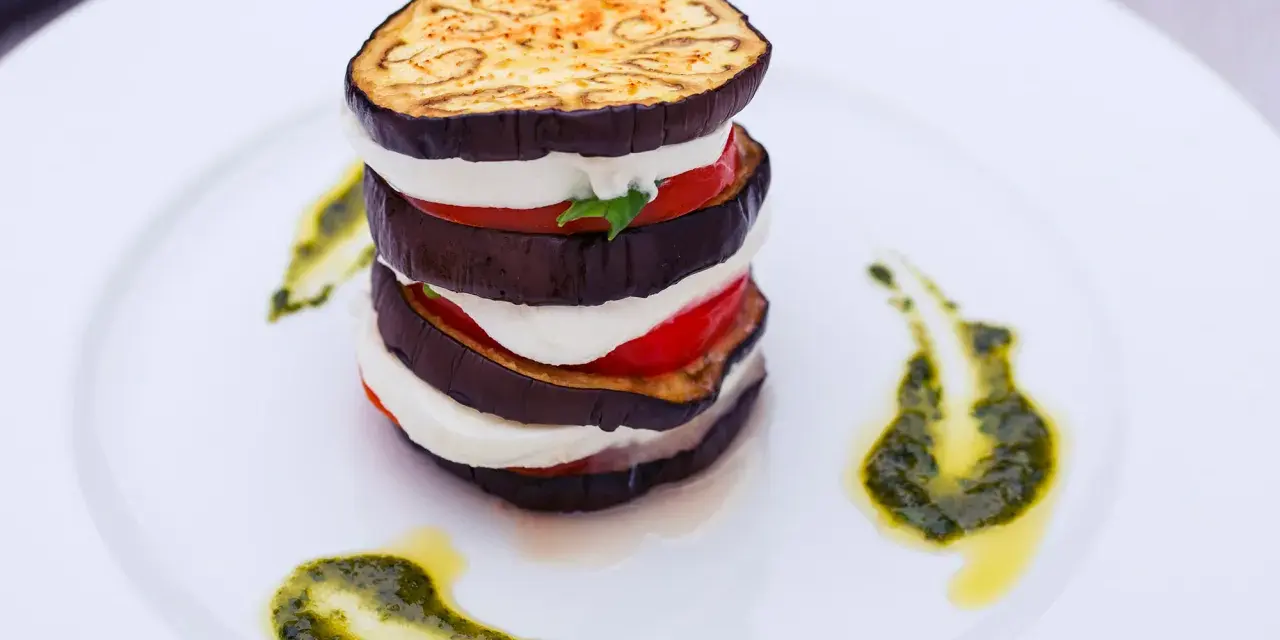 Parmigiana di melanzane a strati, con mozzarella, pomodoro e basilico, guarnita con pesto su piatto bianco.