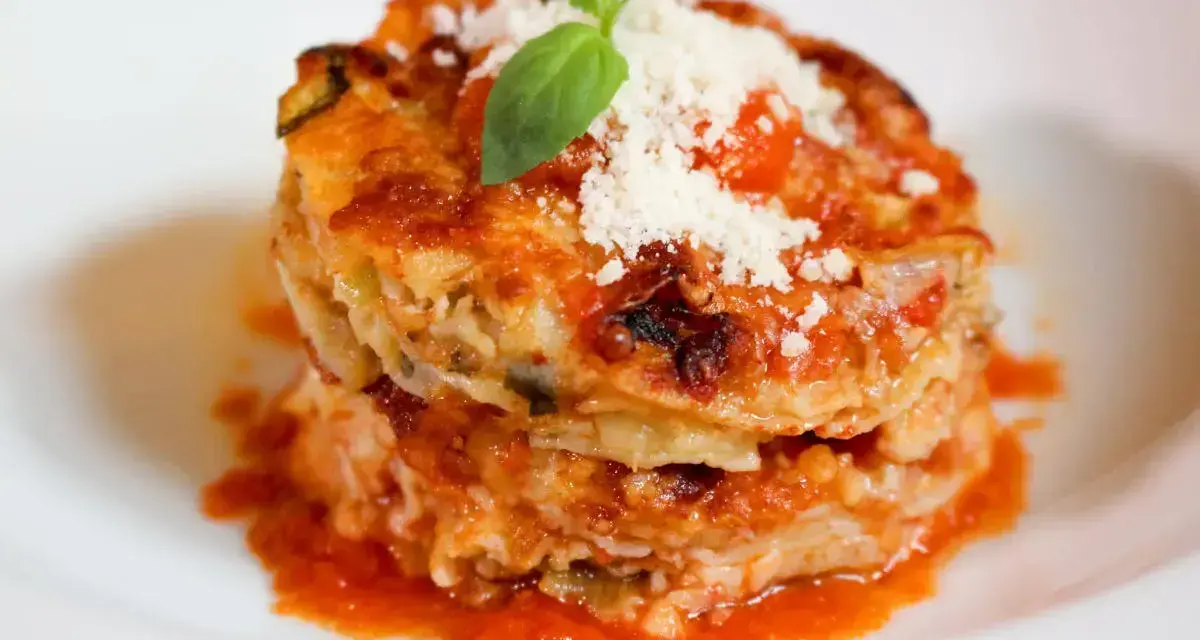Parmigiana di alici, un piatto stratificato con salsa di pomodoro, formaggio e basilico fresco, servito su un piatto bianco.