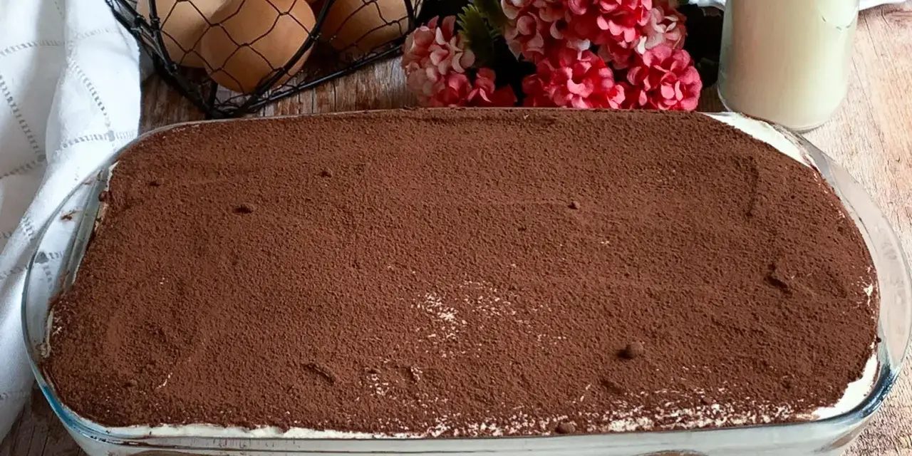 Un invitante tiramisù con panna, cosparso di cacao, pronto per essere gustato. Ingredienti freschi come uova e latte completano la scena.