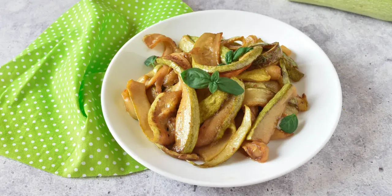 Zucchine in agrodolce, un piatto colorato e saporito, guarnito con basilico fresco su un piatto bianco, accanto a un tovagliolo a pois verdi.