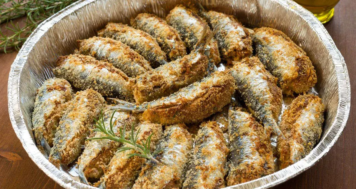 Sardine al forno dorate e croccanti, disposte in una teglia d'alluminio con rametti di rosmarino.