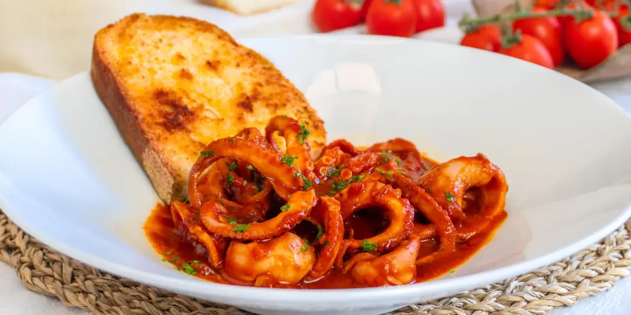Un piatto invitante di moscardini in umido, con tentacoli succulenti in salsa rossa, accompagnati da una fetta di pane tostato dorato.
