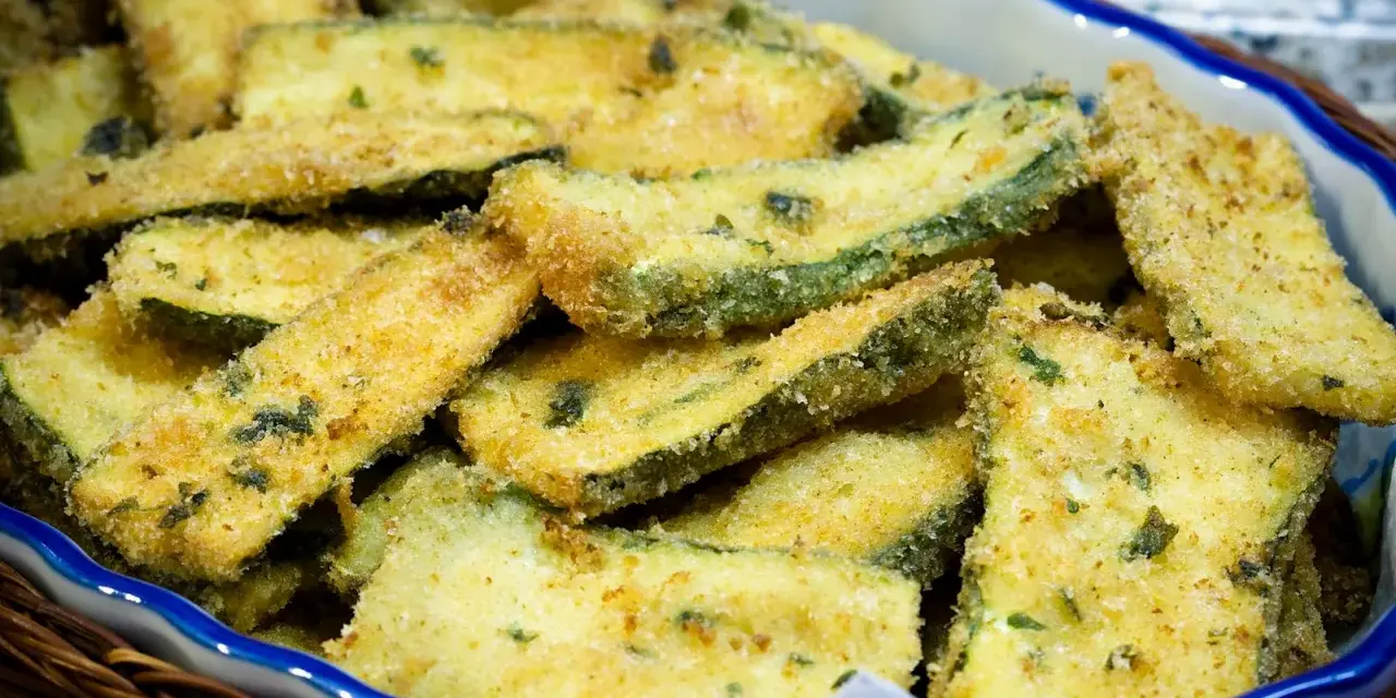 Fette di zucchine gratinate in padella, dorate e croccanti, servite in una ciotola blu bordata di bianco.