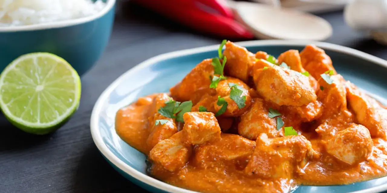 Spezzatino di pollo cremoso e speziato, guarnito con prezzemolo fresco, servito su un piatto blu con riso bianco e spicchio di lime.