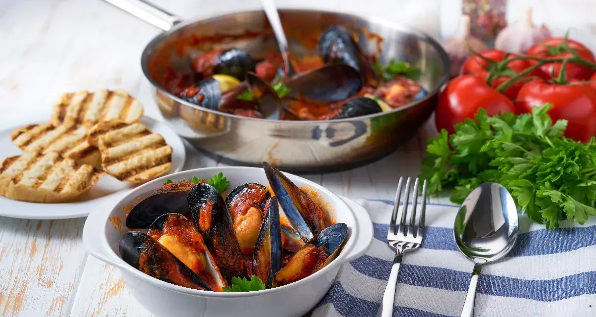 Cozze alla tarantina fumanti in una ciotola bianca, accompagnate da pane grigliato e ingredienti freschi.