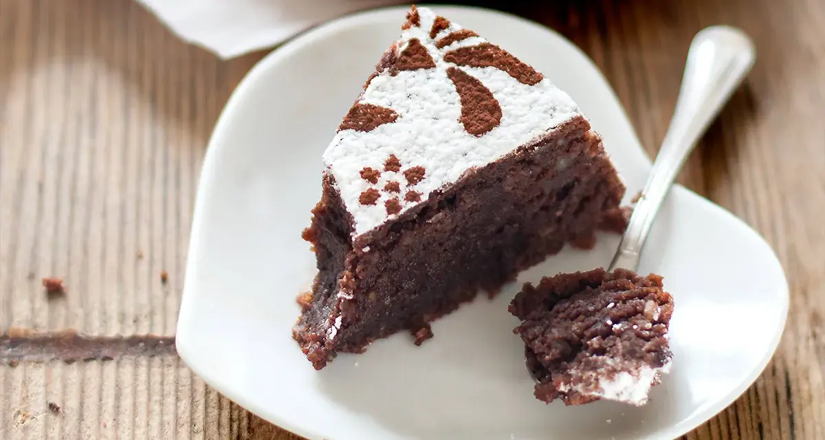 Una fetta di torta caprese ricetta originale napoletana, cosparsa di zucchero a velo e cacao, su un piattino bianco a forma di cuore.