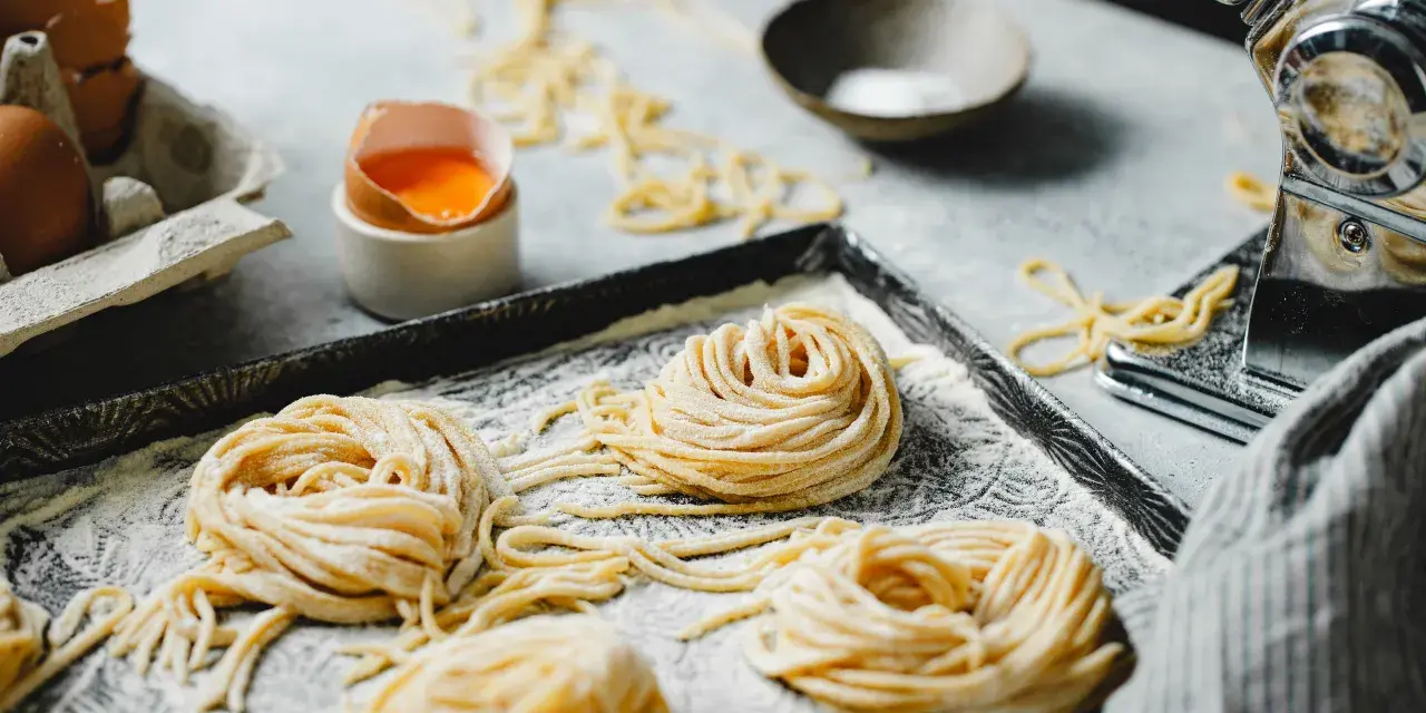 Nidi di pasta fatta in casa su un vassoio infarinato, con uova e una macchina per la pasta sullo sfondo.