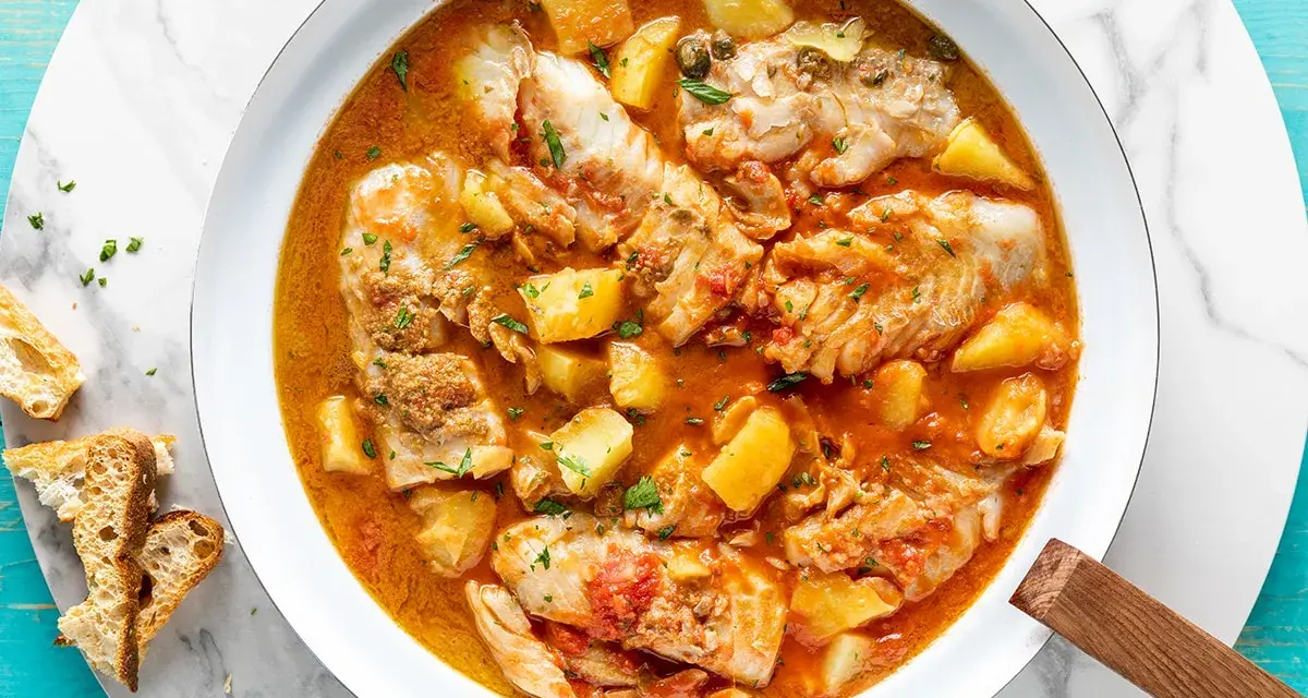 Un saporito merluzzo in umido con patate e pomodoro, guarnito con prezzemolo fresco e accompagnato da pane tostato.