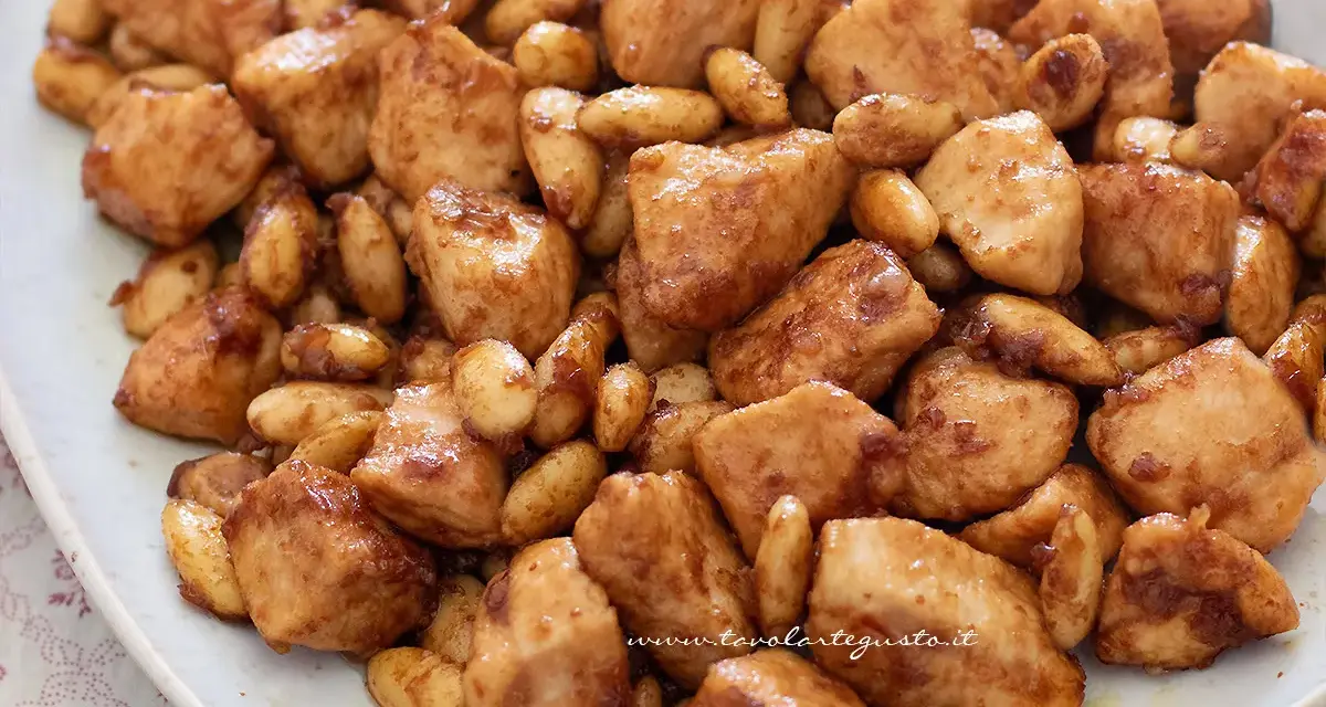 Close-up of bite-sized chicken pieces and almonds coated in a glossy brown sauce on a white plate.