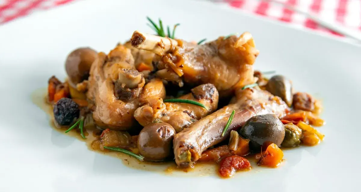 Coniglio alla cacciatora ricetta della nonna: succulenti pezzi di coniglio con olive, pomodorini e rosmarino su un piatto bianco.