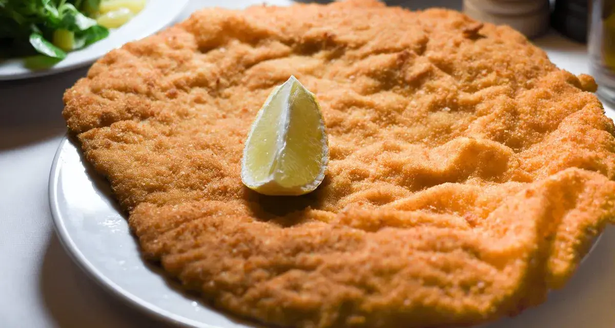 Una cotoletta dorata, grande come un'orecchia di elefante, servita con una fetta di limone e un'insalata fresca.