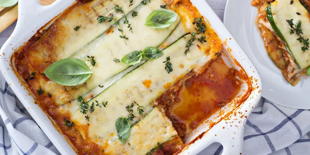 Lasagne di zucchine al forno, dorate e filanti, guarnite con basilico e timo. Una porzione è già servita su un piatto bianco.