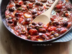 Un cucchiaio di legno raccoglie un sugo ricco e saporito, pronto per condire la pasta alla puttanesca. Pomodoro, olive e capperi si mescolano in un tripudio di sapori mediterranei.