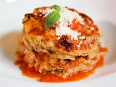 Parmigiana di alici, un piatto stratificato con salsa di pomodoro, formaggio e basilico fresco, servito su un piatto bianco.