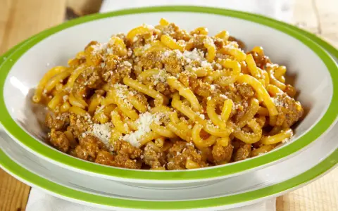 Piatto di gramigna alla salsiccia, condita con ragù e parmigiano grattugiato, servita in una ciotola con bordo verde.