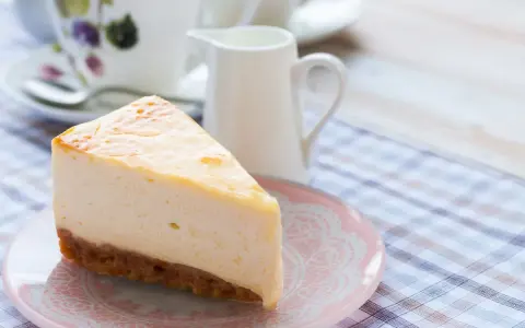 Una fetta di cheesecake con mascarpone, cremosa e invitante, su un piattino rosa. Sullo sfondo, una tazza da tè e una lattiera.