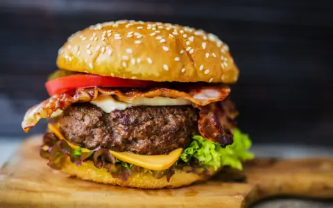 Hamburger fatti in casa con bacon croccante, formaggio filante, pomodoro fresco e lattuga su un tagliere di legno.