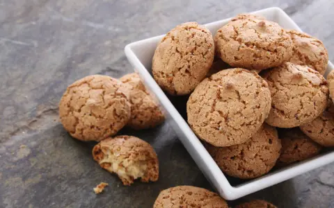 Un mucchio di croccanti biscotti alle mandorle in una ciotola bianca, alcuni sparsi su una superficie grigia.