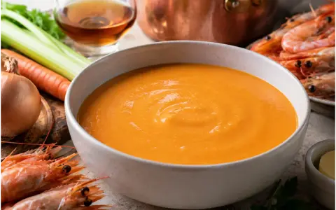 Una ciotola di cremosa bisque di gamberi, con gamberi freschi, carote, sedano e cipolla sullo sfondo.