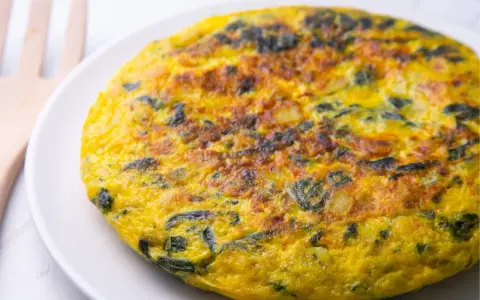 Frittata di spinaci dorata e invitante su un piatto bianco, con un utensile in legno sullo sfondo.