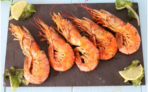 Cinque succulenti gamberoni alla griglia, serviti su una pietra scura con spicchi di limone e insalata fresca.