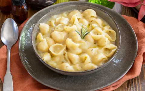 Una ciotola di minestra di patate cremosa con pasta a conchiglia, guarnita con rosmarino. Un piatto confortante e saporito.