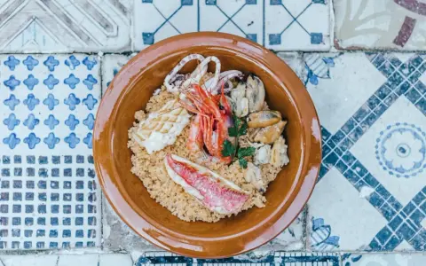 Un invitante cous cous di pesce con gamberi, calamari, cozze e pesce rosso, guarnito con prezzemolo, servito in una ciotola di terracotta su un piatto di maioliche.