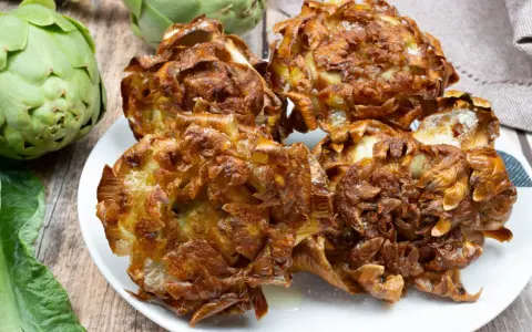 Carciofi alla giudia dorati e croccanti su un piatto bianco, con carciofi freschi sullo sfondo.