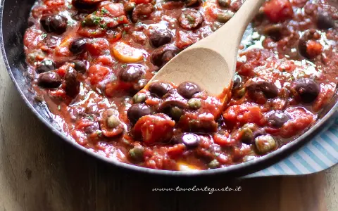 Un cucchiaio di legno raccoglie un sugo ricco e saporito, pronto per condire la pasta alla puttanesca. Pomodoro, olive e capperi si mescolano in un tripudio di sapori mediterranei.
